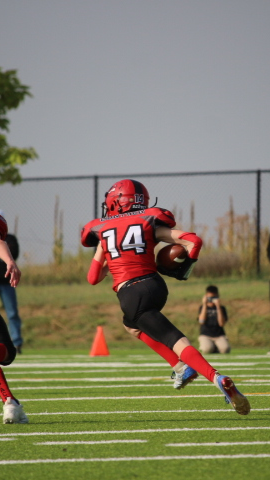 Football players in action on a grassy field; one runs with the ball, pursued by two opponents.