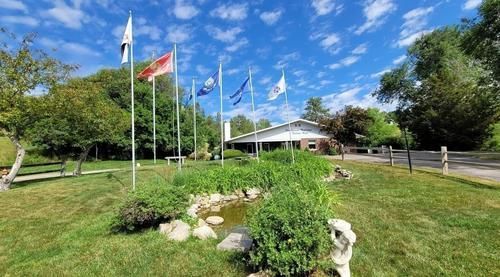 Flags fly in front of a white building with a small pond. Green grass and blue sky.