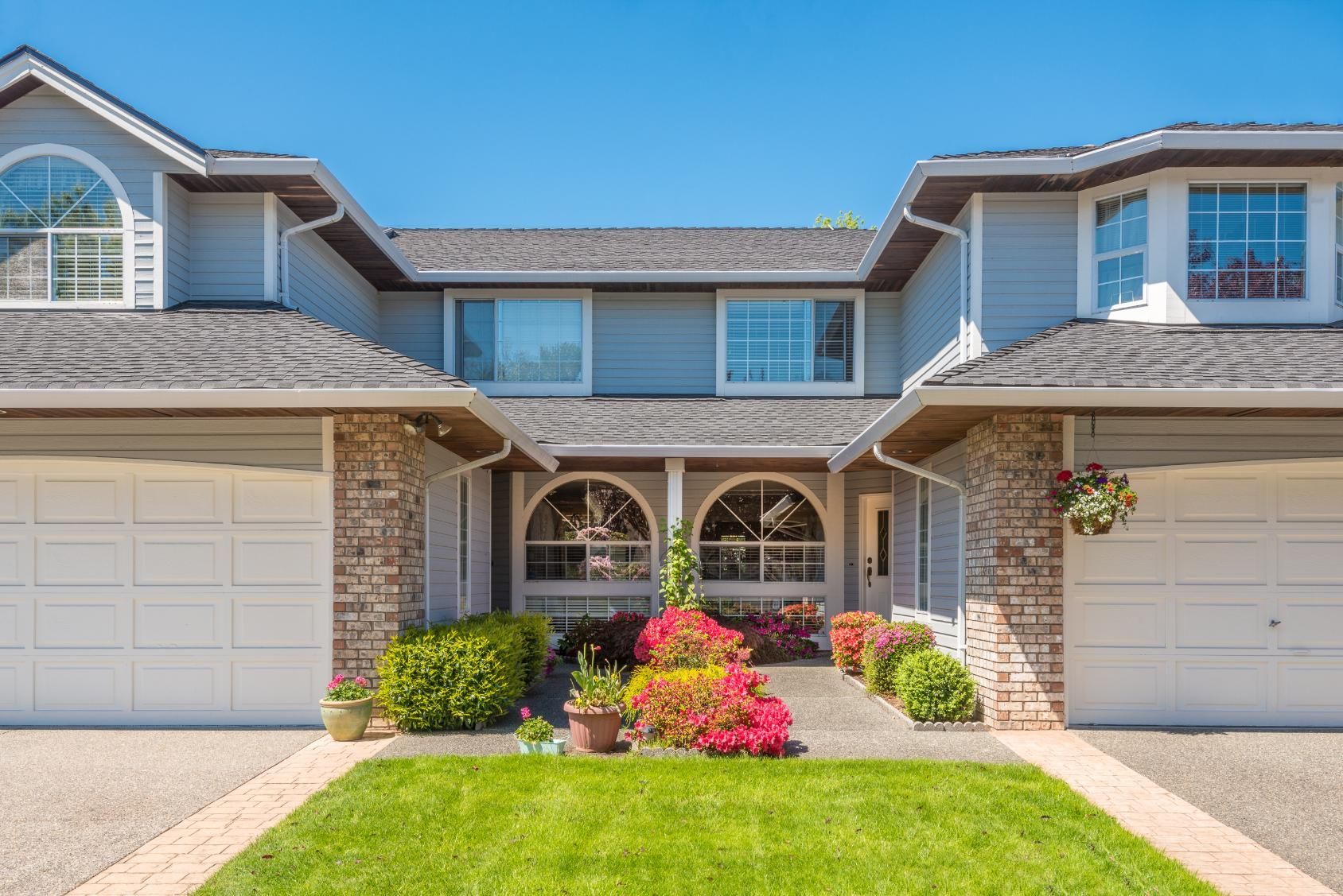 Two-story gray townhomes with white garage doors and a walkway lined with flowers and shrubs under a blue sky.