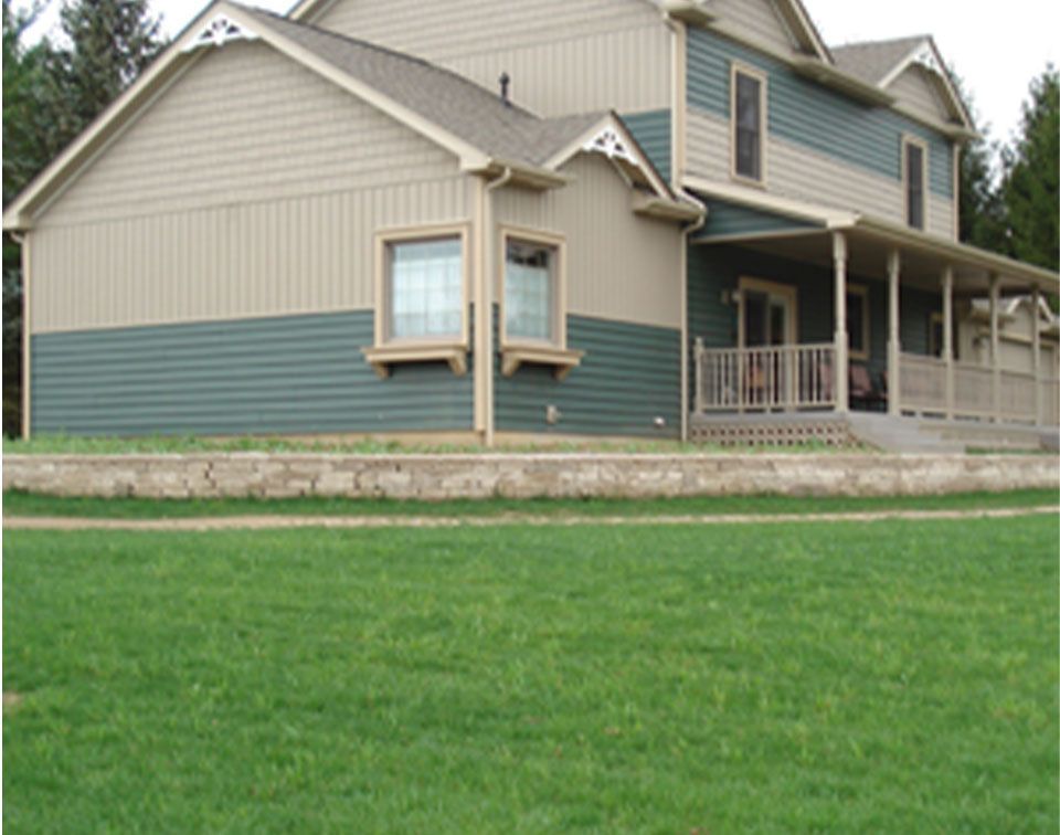 Two-story house with green and tan siding, a porch, and a stone retaining wall.