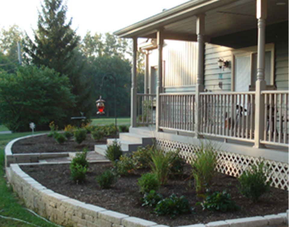A house with a porch and flowerbeds, brown retaining wall, with a bird feeder in the background.