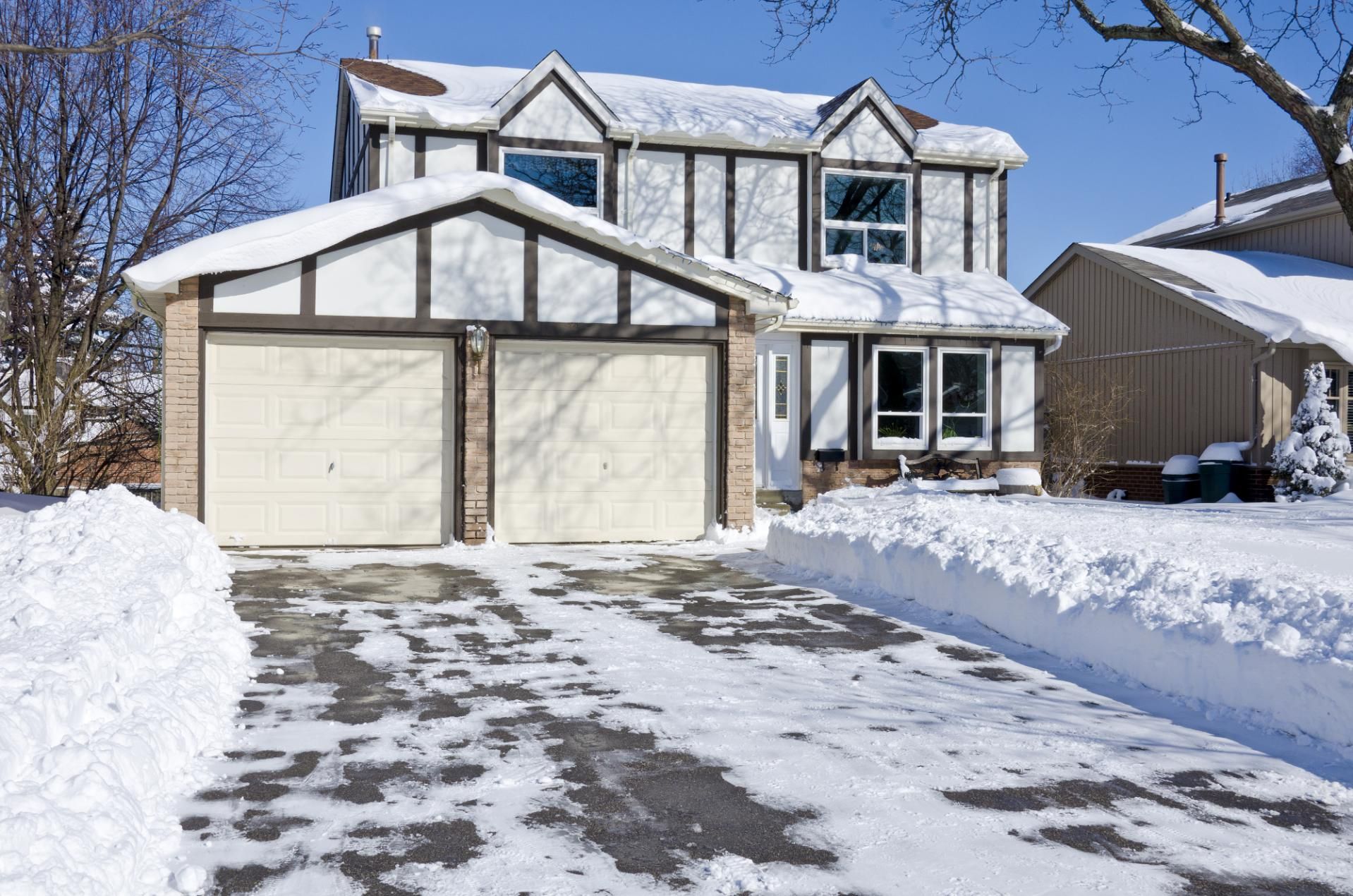 Two-story house with a snow-covered yard and driveway, a detached garage with two bays, and a sunny sky.