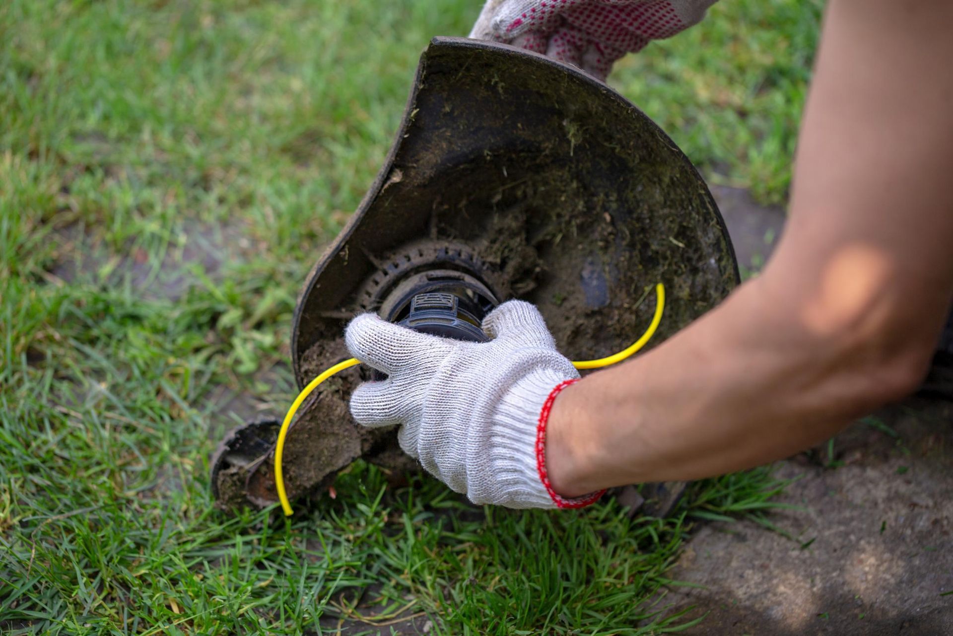 Person wearing gloves, replacing trimmer line on a weed eater, outdoors on grass.