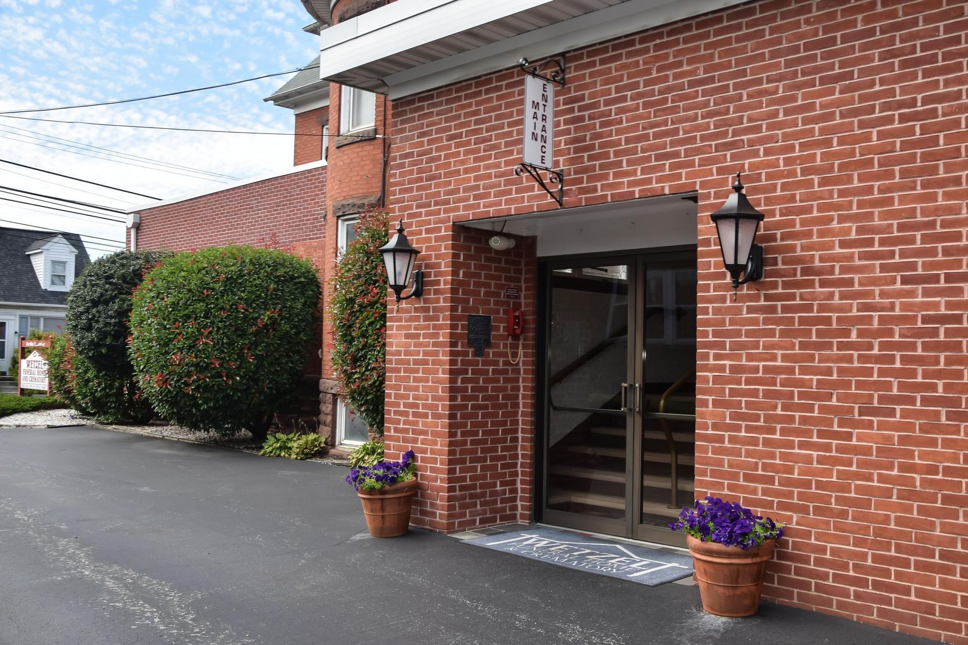 Brick building entrance with glass doors, wall lanterns, and flower pots beside a paved driveway.