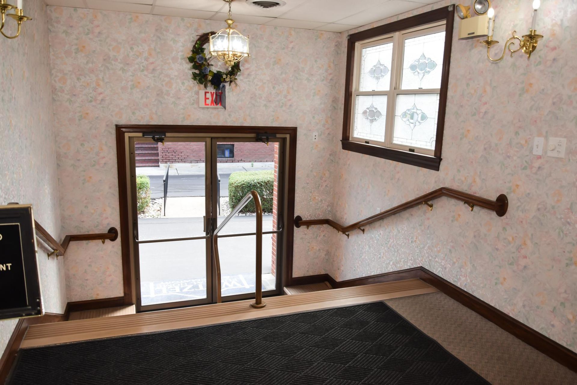 Apartment entryway with glass double doors, staircase railings, patterned wallpaper, and a dark floor mat.