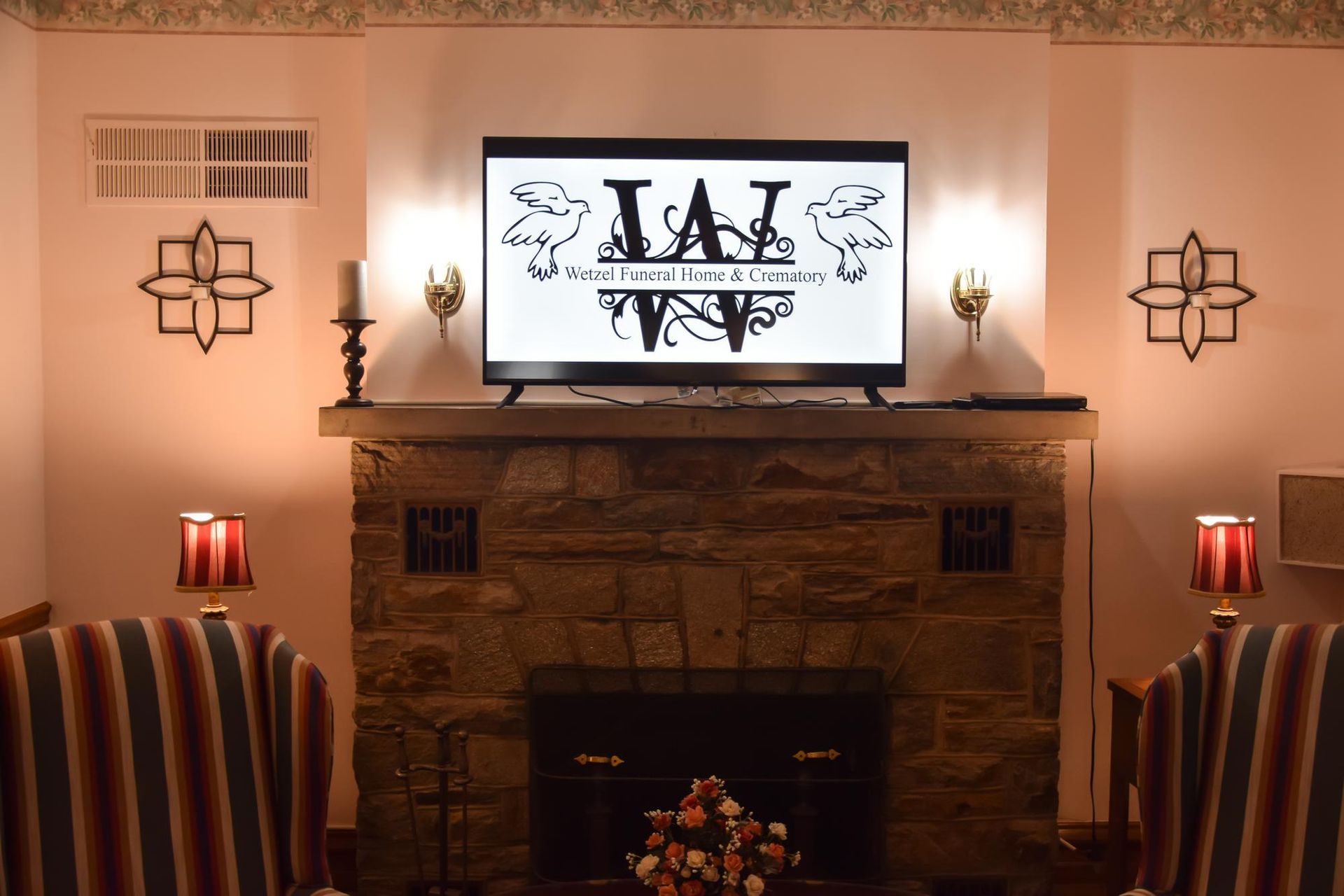 Living room fireplace with a TV displaying the “WI” logo, warm lamps, and striped chairs