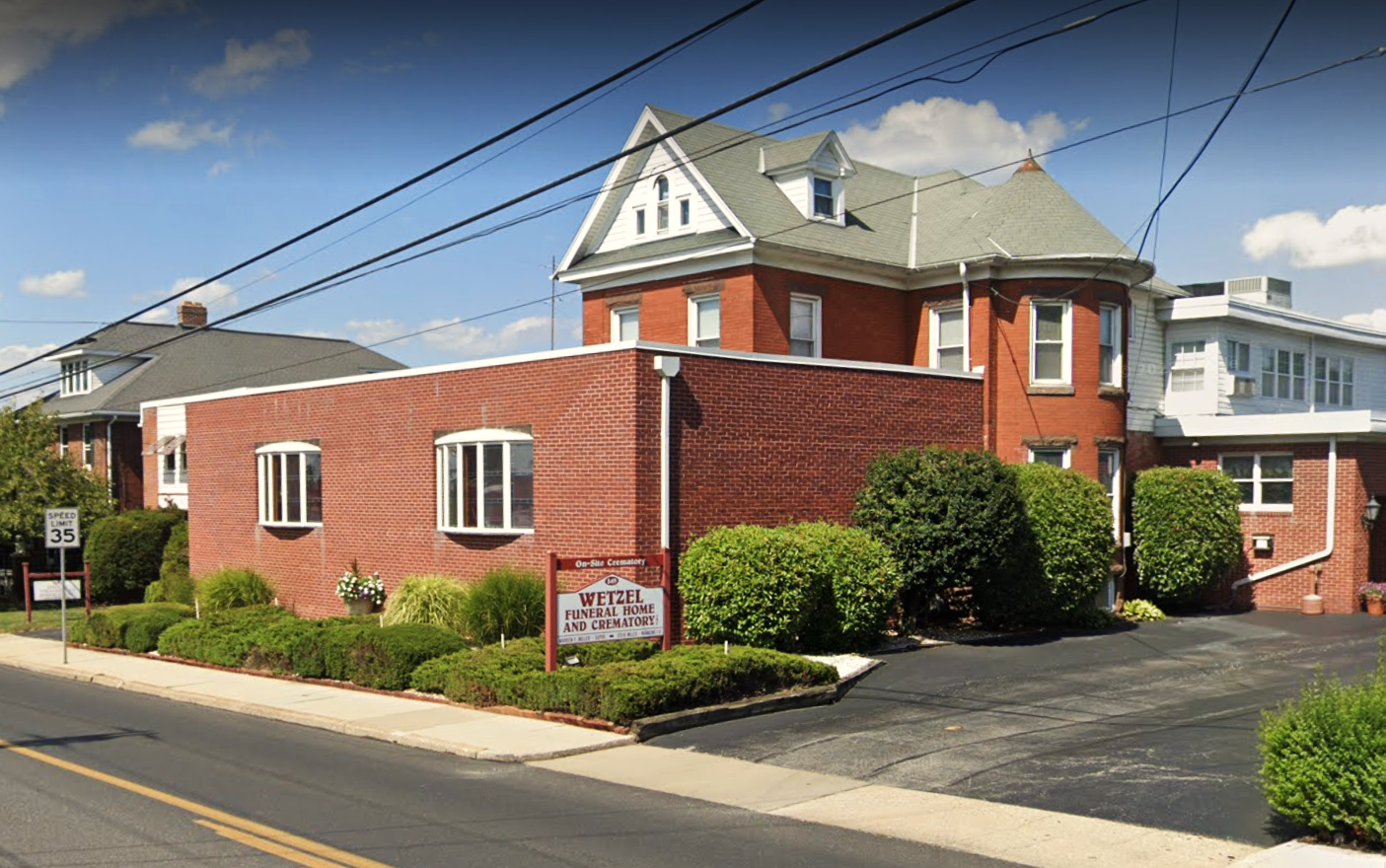 Red brick church building with a steep roof, driveway, and sign beside a suburban street