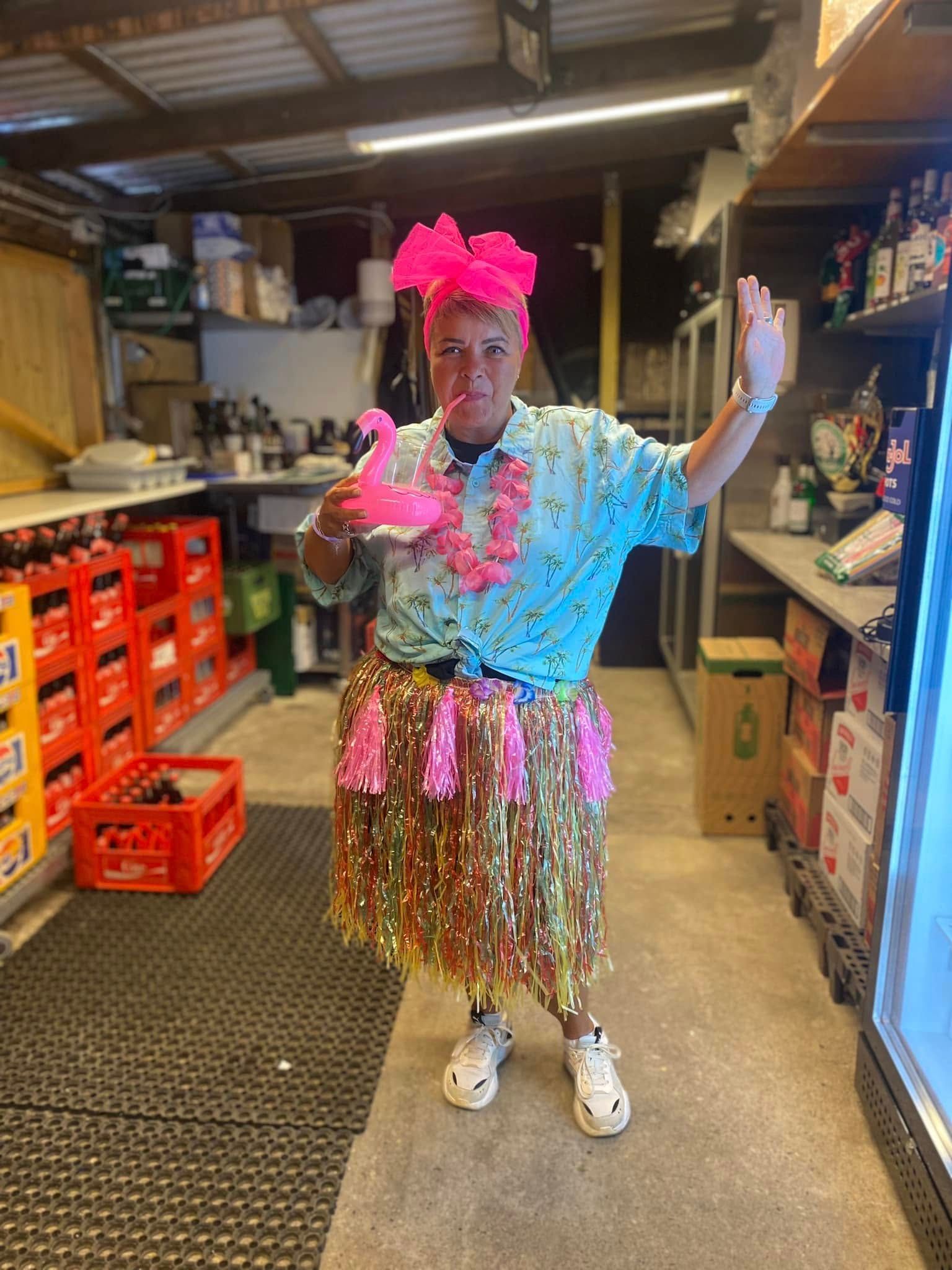 En mand iført hula-nederdel og lyserødt pandebånd står i et værelse.