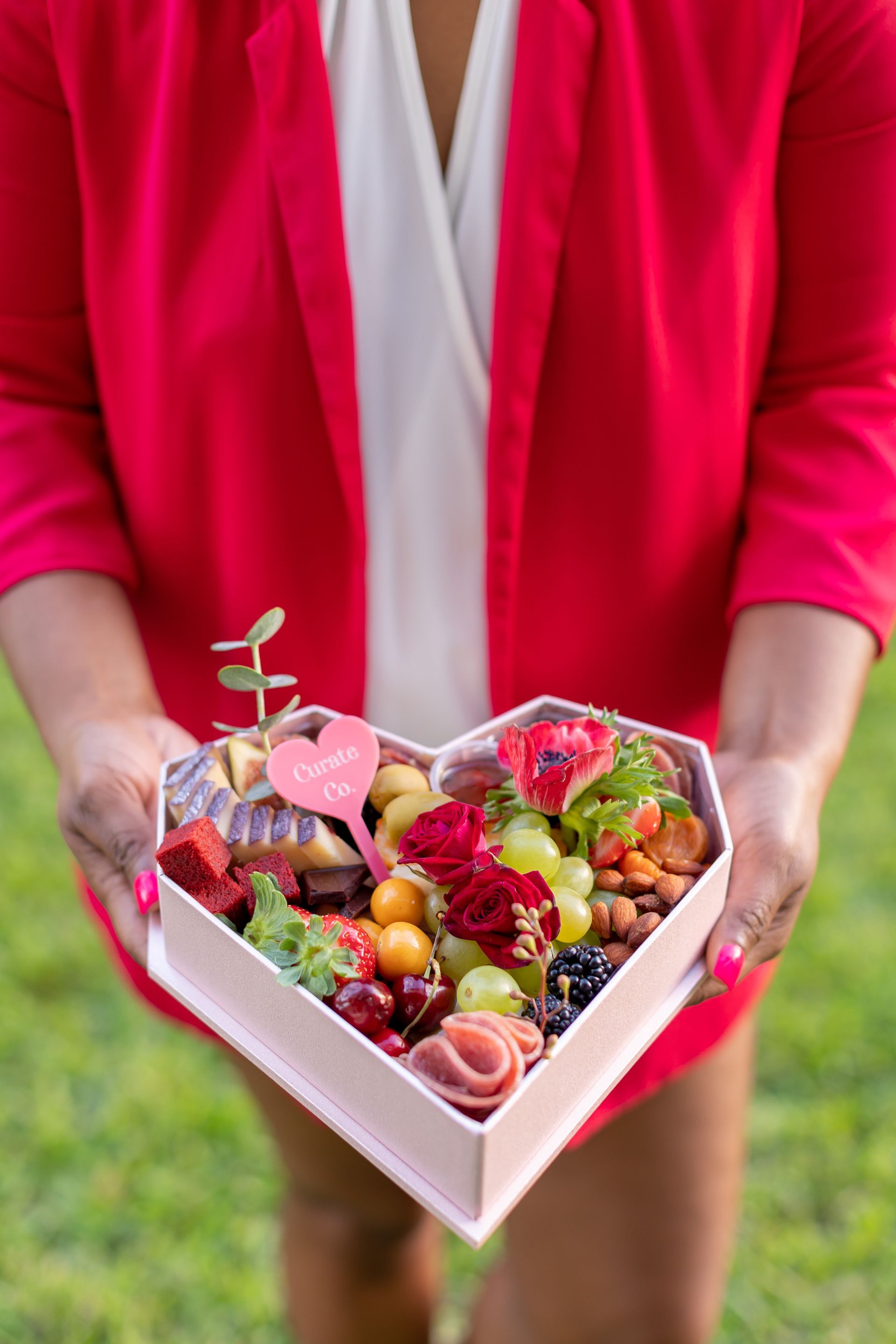 A woman is holding a heart shaped box filled with fruits and flowers.