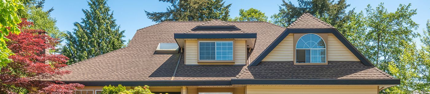 House with brown shingle roof, dormers, and trees under a clear blue sky.
