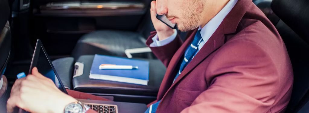A businessman in a car on a phone call works on a laptop. He wears a suit and watch.