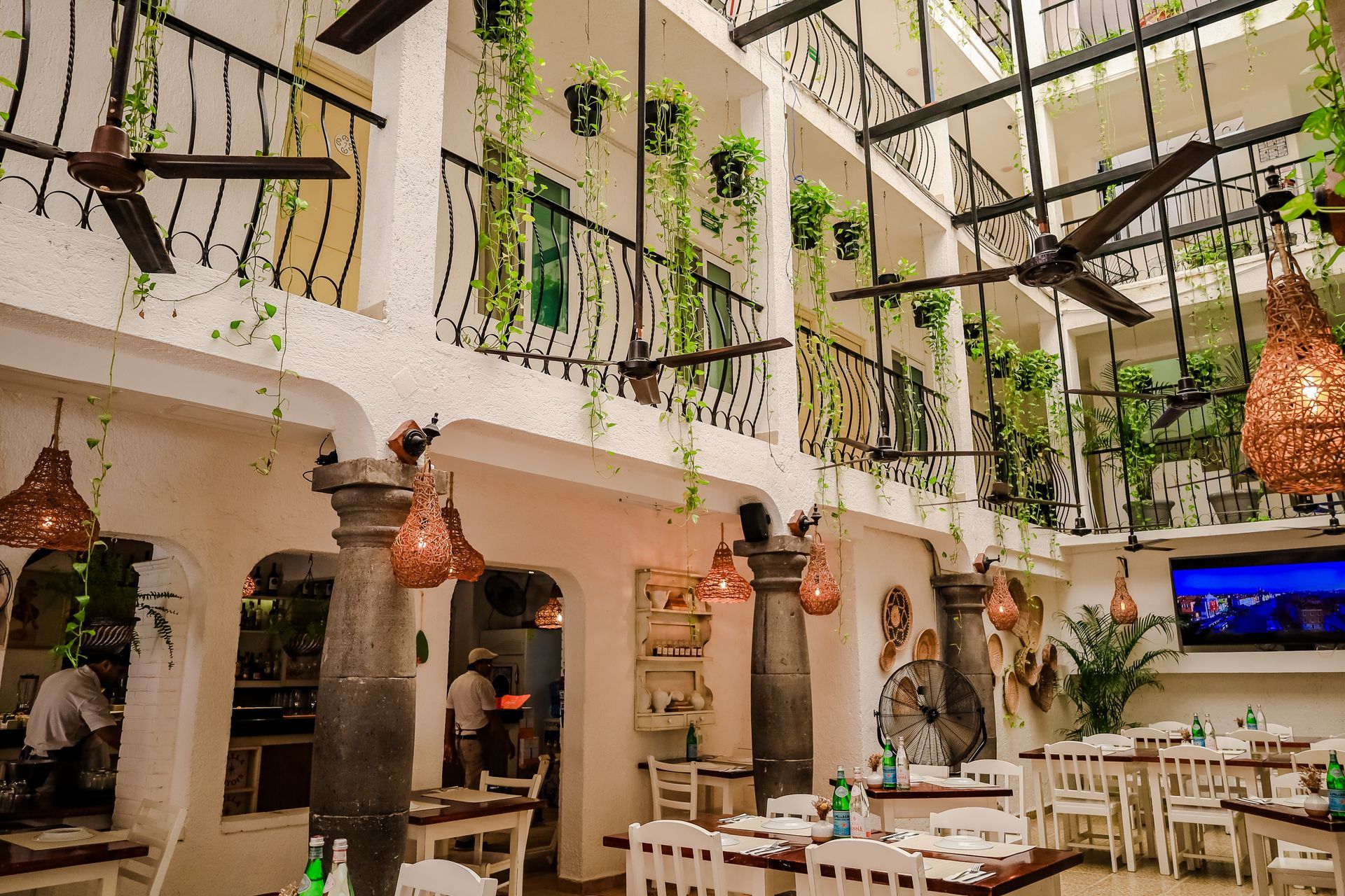 Restaurant interior with white walls, balconies, hanging plants, and tables set for dining.