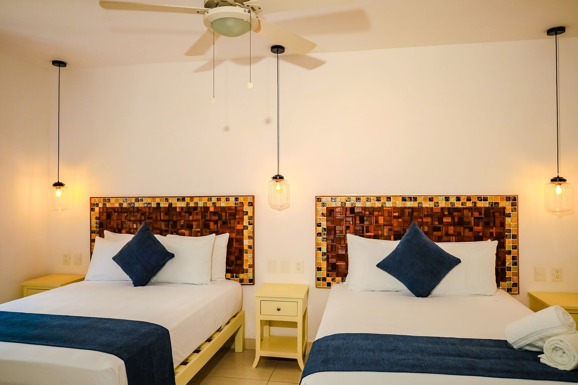 Two beds in a hotel room, with decorative headboards, nightstands, and hanging lights.