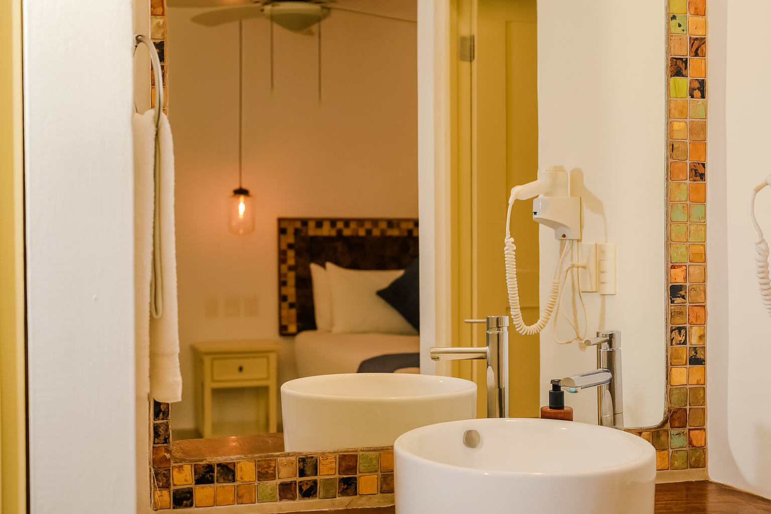 Bathroom mirror reflecting a bedroom: white sink, mosaic trim, towel, bed, nightstand, and hanging light.