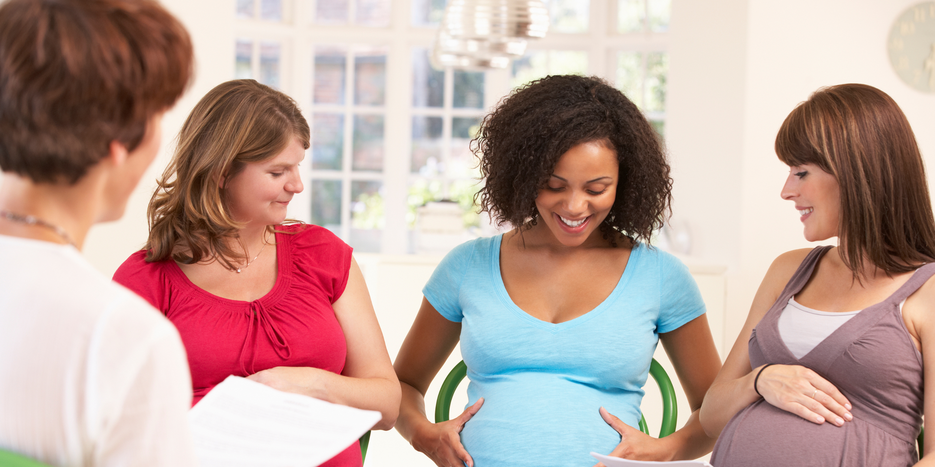 Four pregnant women in a group, touching bellies, smiling, indoors.
