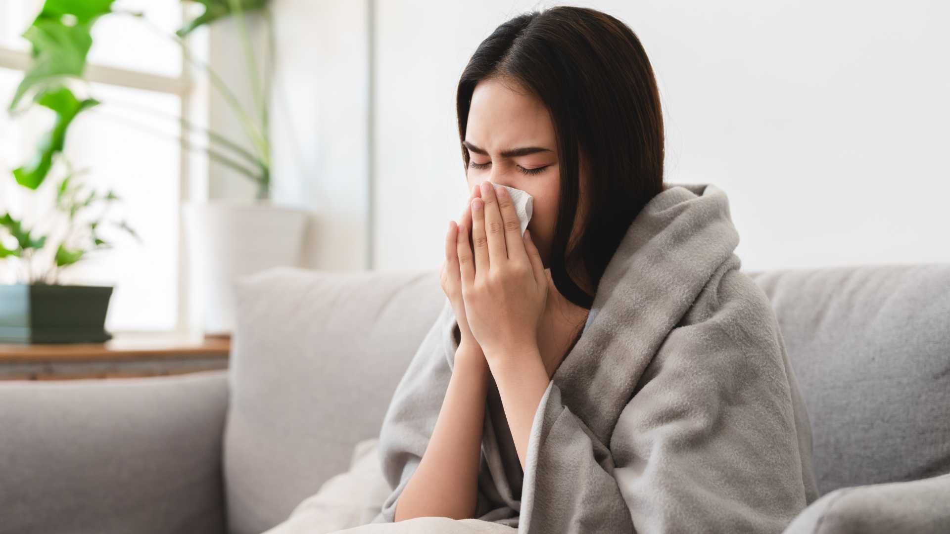 A woman is sitting on a couch blowing her nose into a napkin.