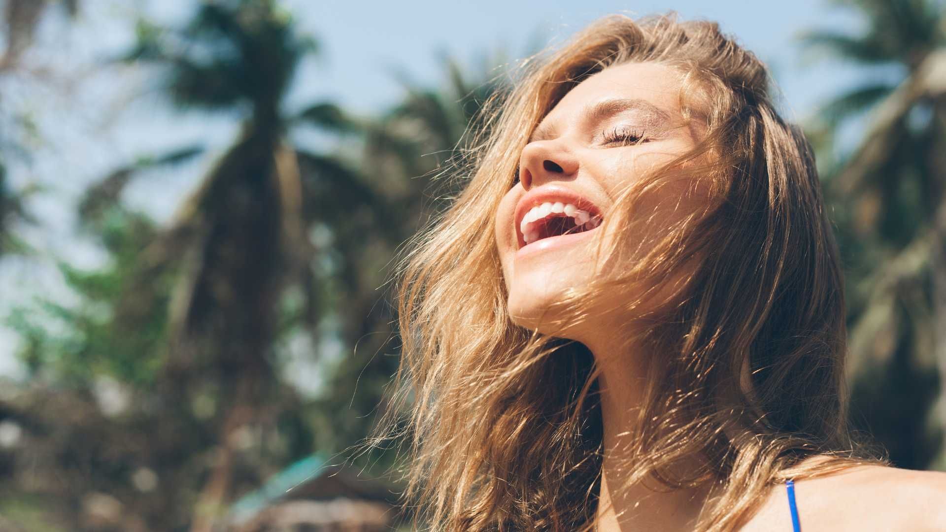 A woman is laughing on the beach with palm trees in the background.