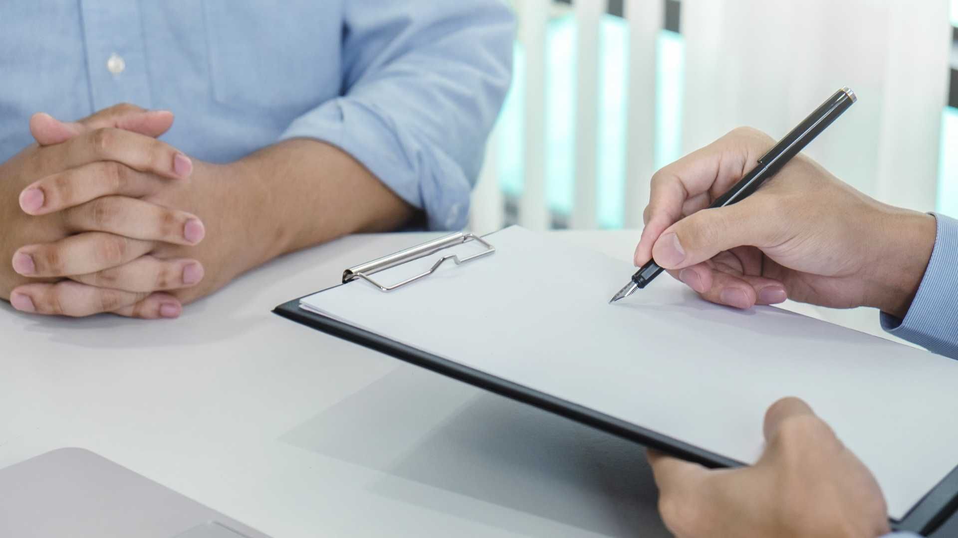 A man is sitting at a table while another man writes on a clipboard.