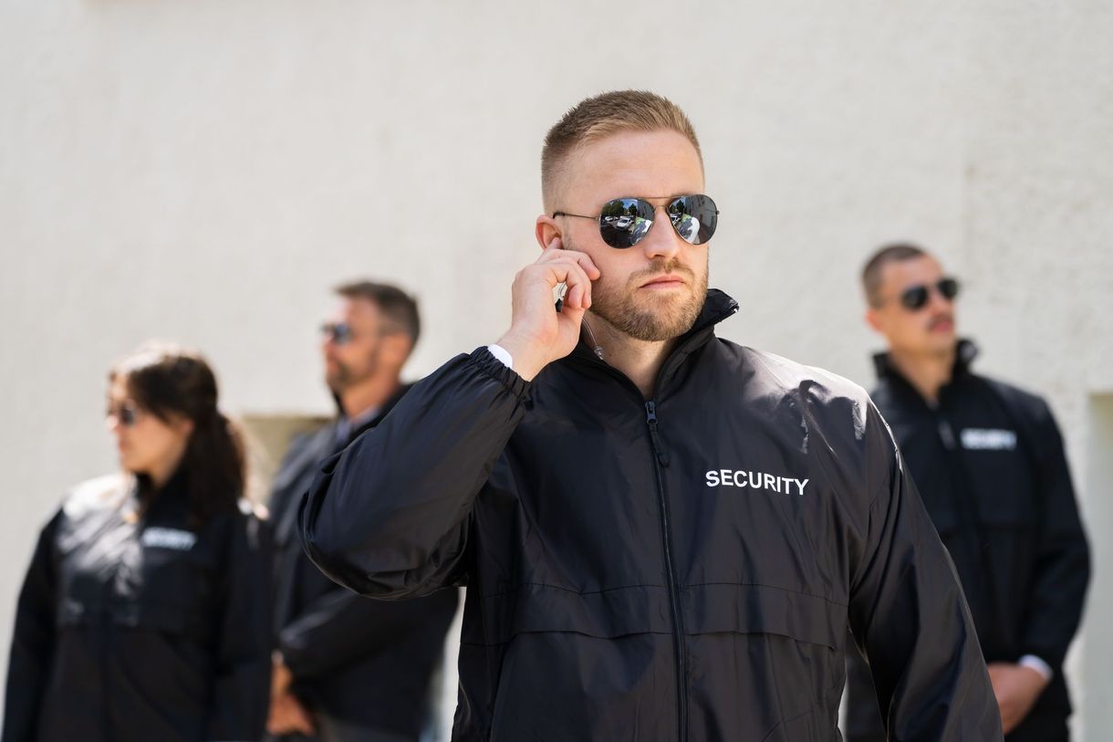 Man in black jacket and sunglasses touching his ear, with two blurred people standing behind him outdoors.