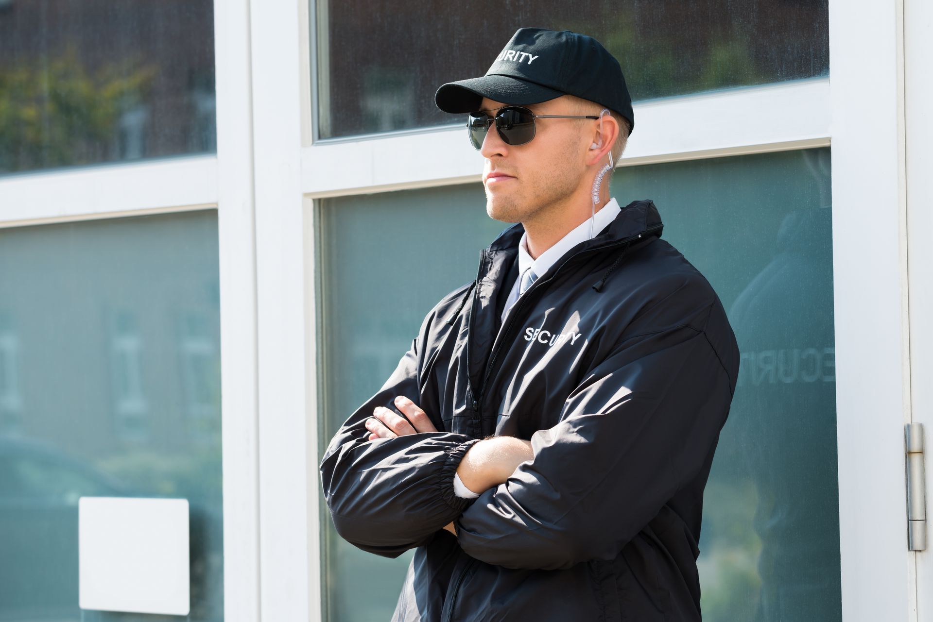 Man in black jacket and cap with sunglasses, standing with arms crossed outside a building Man in black jacket and cap with sunglasses, standing with arms crossed outside a building