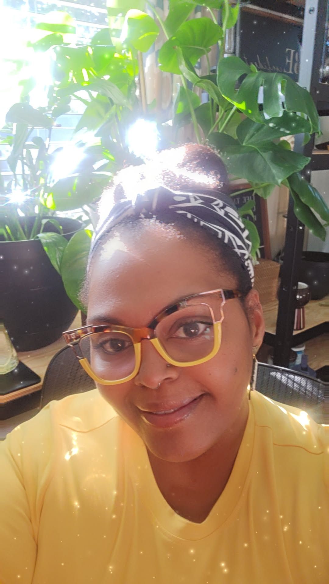 Black woman wearing a yellow top and yellow and tortoise shell glasses, a black and white headband around her natural hair in a bun. She is seated, smiling with natural sunlight behind her and many green plants in pots.