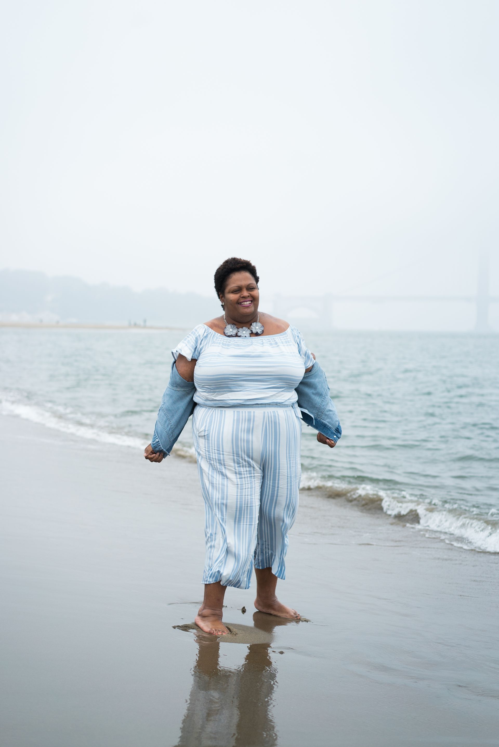 Woman in a light blue striped outfit standing barefoot on a beach by the ocean