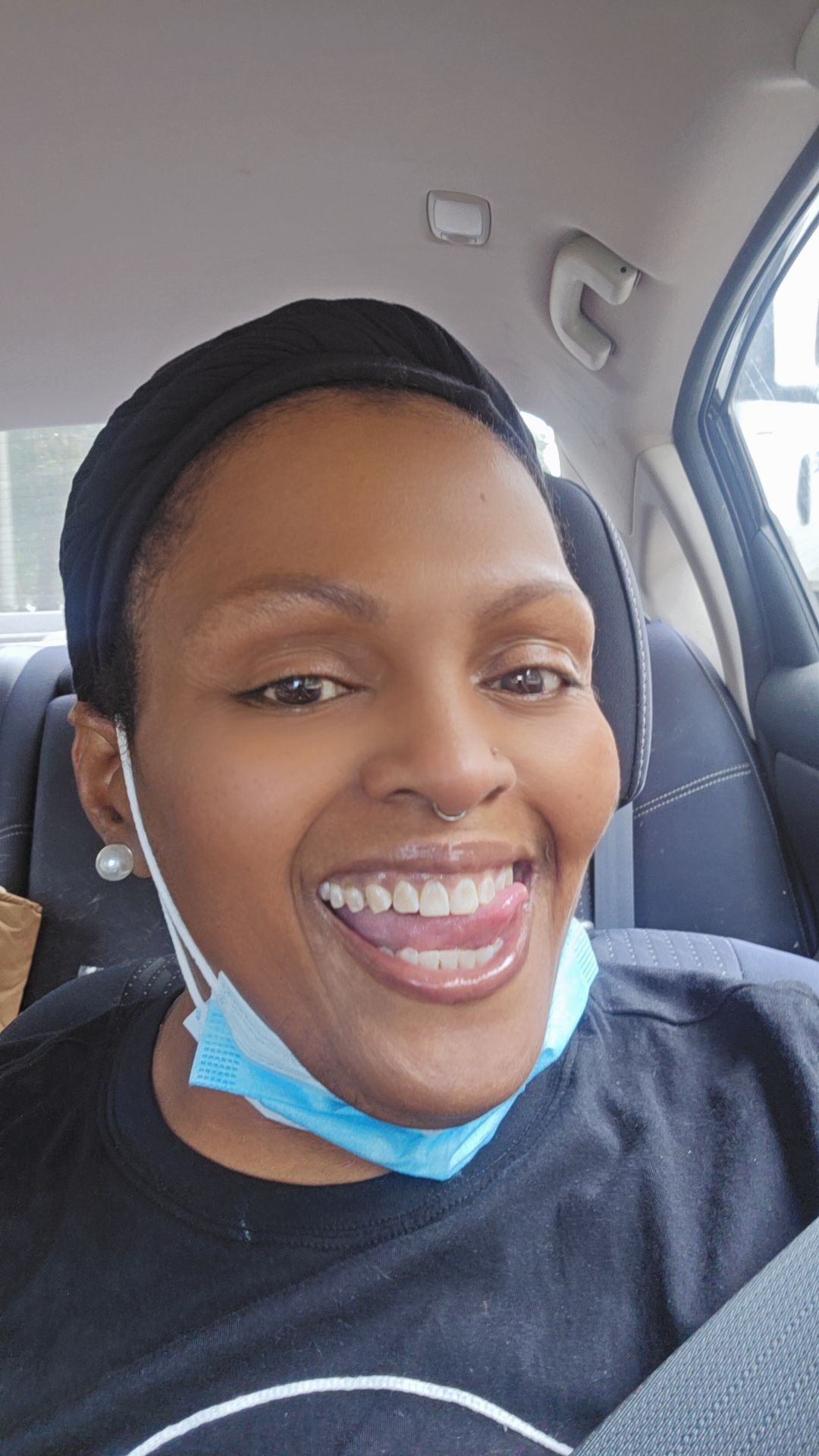 Black Woman wearing a blue mark under her chin smiling with her tongue sticking out to the right. Pearl earrings and a black head covering. 
