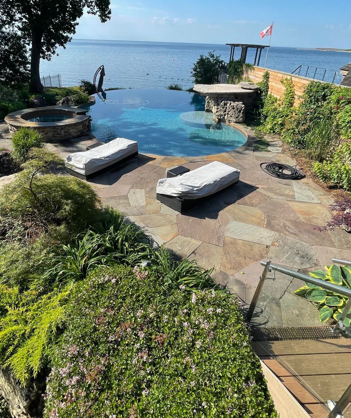 Stone patio with pool and lounge chairs overlooking a tranquil body of water under a blue sky.