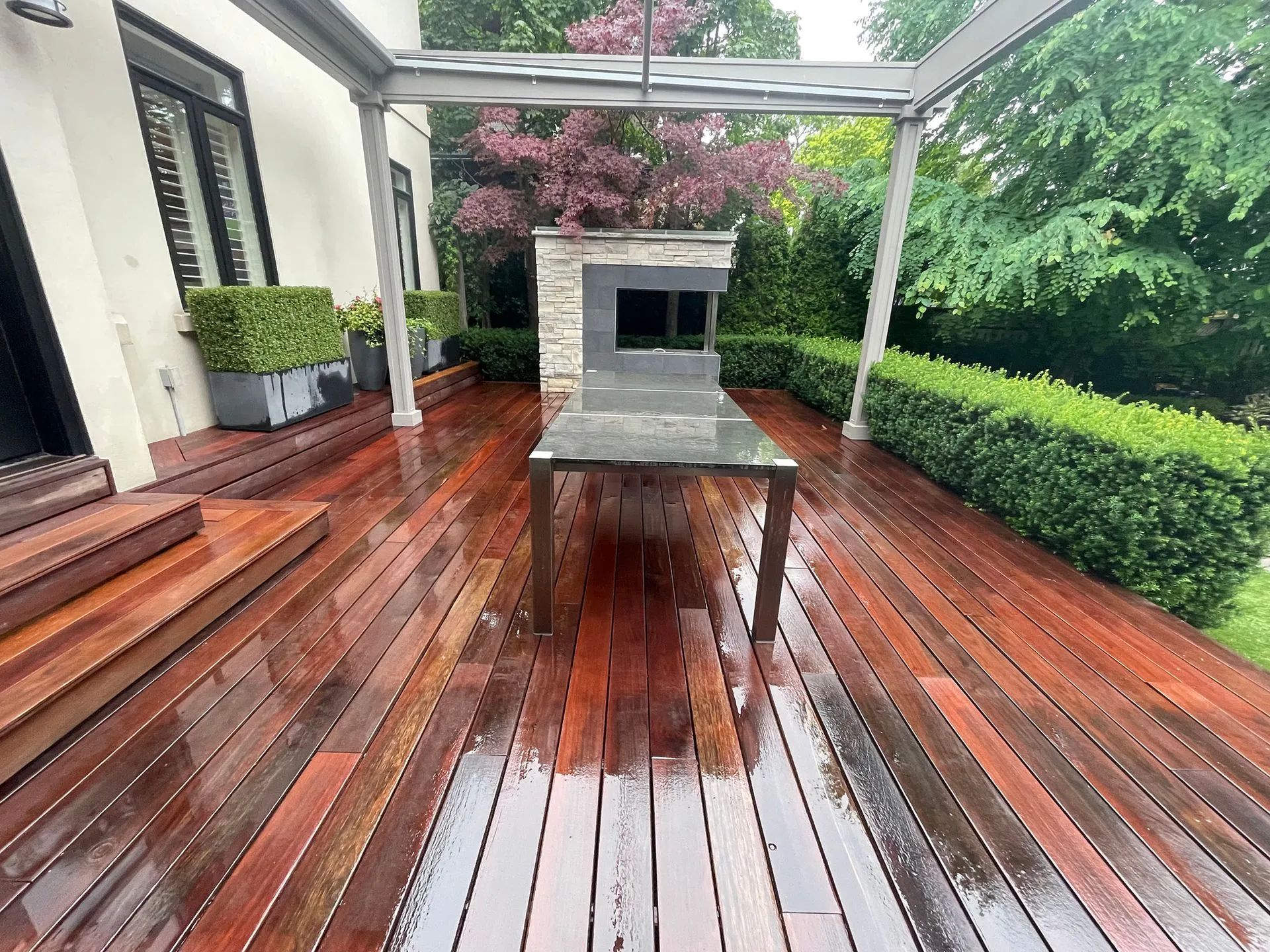 Wooden deck with a table, an outdoor fireplace, and trimmed hedges. Covered by a canopy.