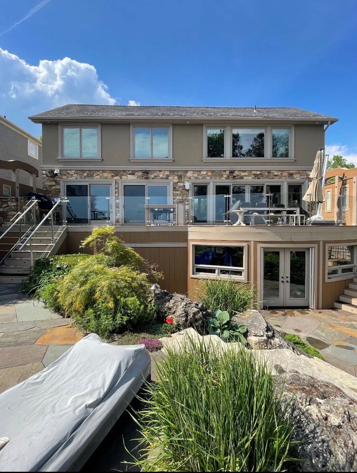 Two-story house with stone facade, overlooking water. Blue sky, deck, boat covered.