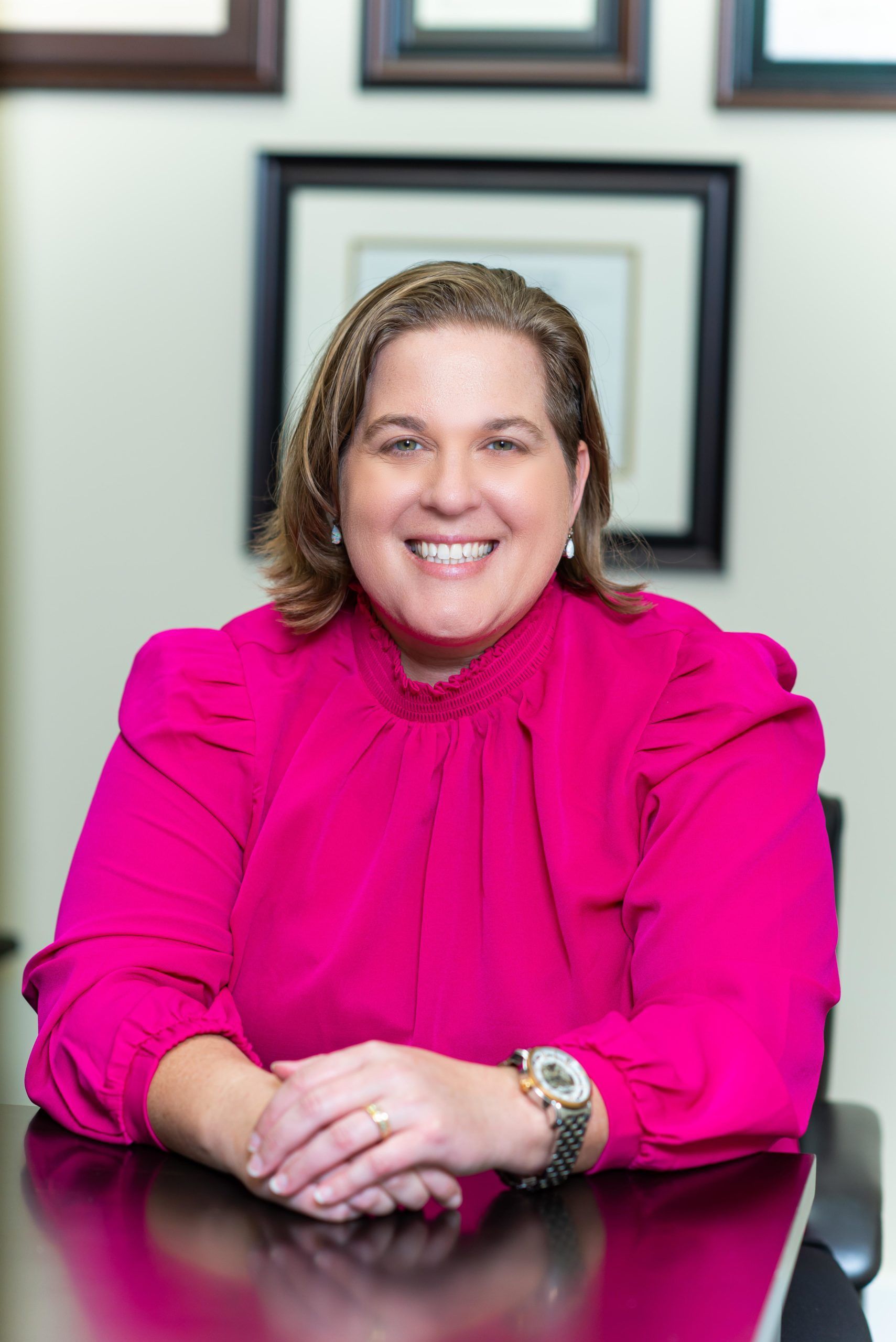 A woman in a pink shirt is sitting at a table with her hands folded and smiling.