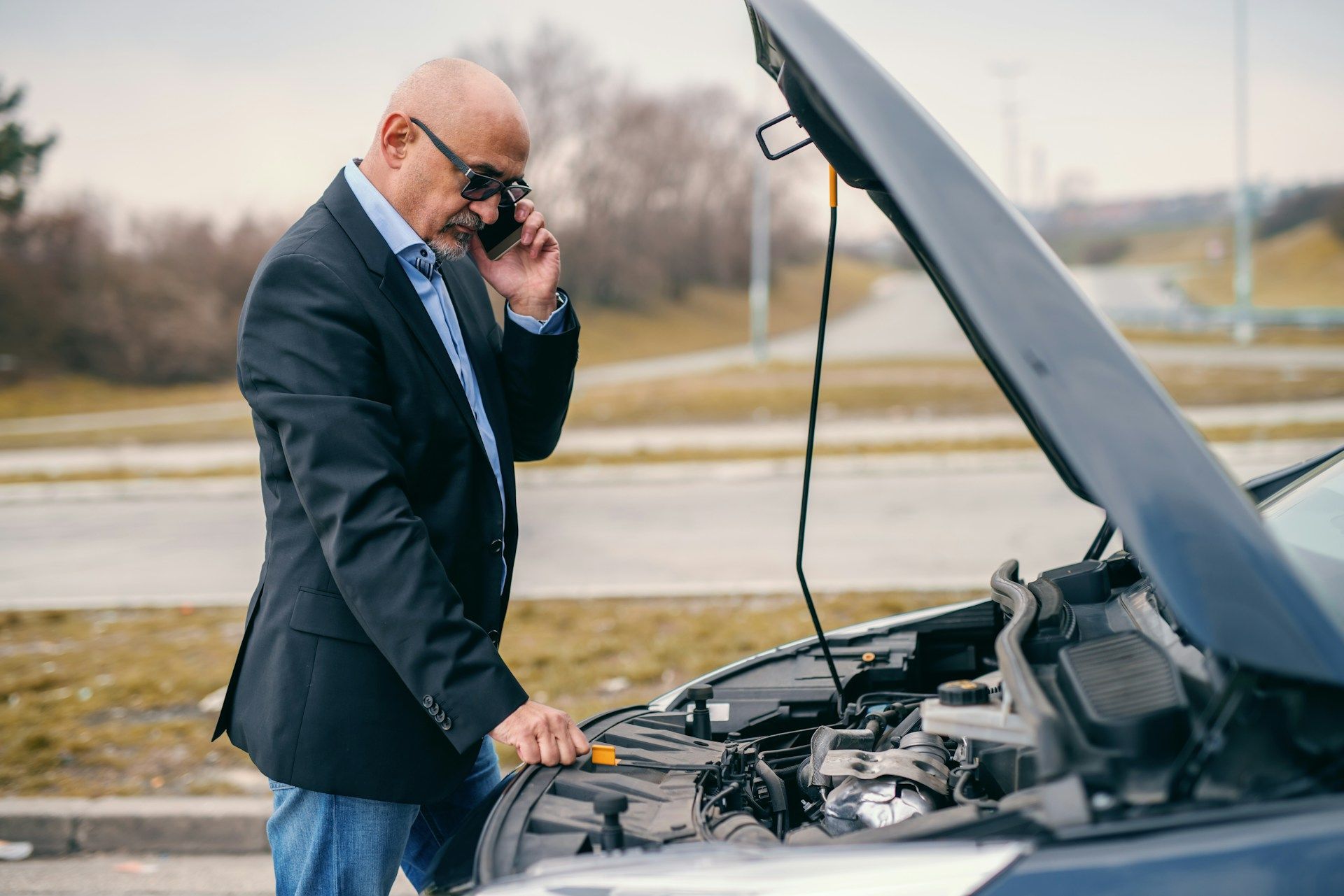A man is talking on a cell phone while looking under the hood of his broken down car.