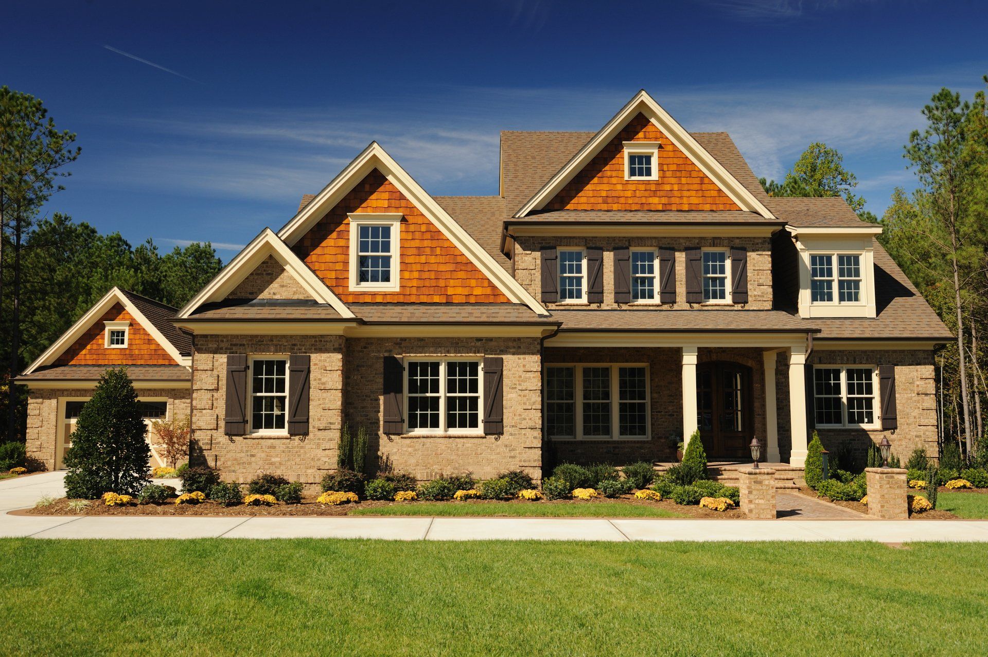 Large tan stone suburban house with orange gables, a front porch, and a manicured lawn under a blue sky