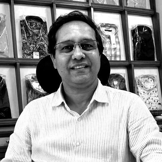 A man wearing glasses and a striped shirt is sitting in front of a wall of shirts.