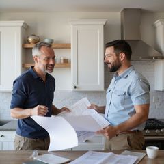 Two men reviewing blueprints in a kitchen with white cabinets, smiling and discussing plans.