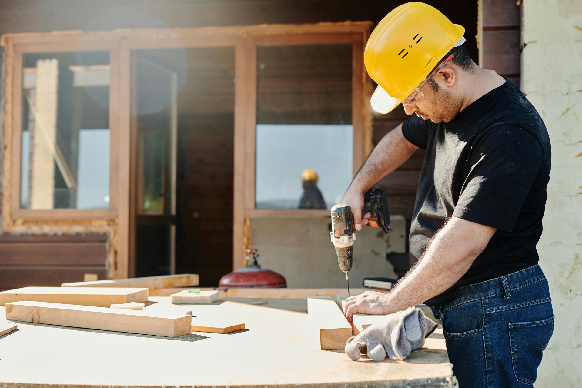 Person wearing a yellow hard hat drills wood on a table outdoors near a building.