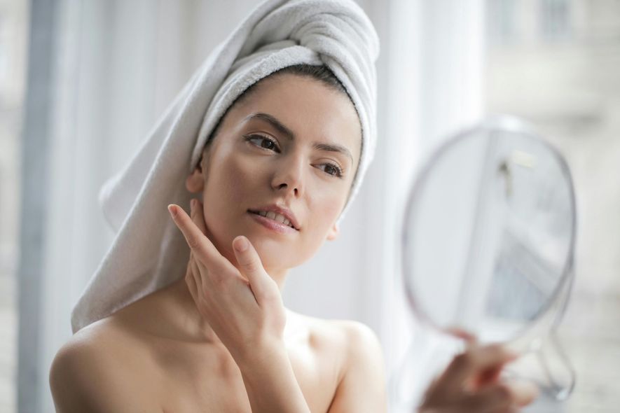 A woman with a towel wrapped around her head is looking at her face in a mirror.