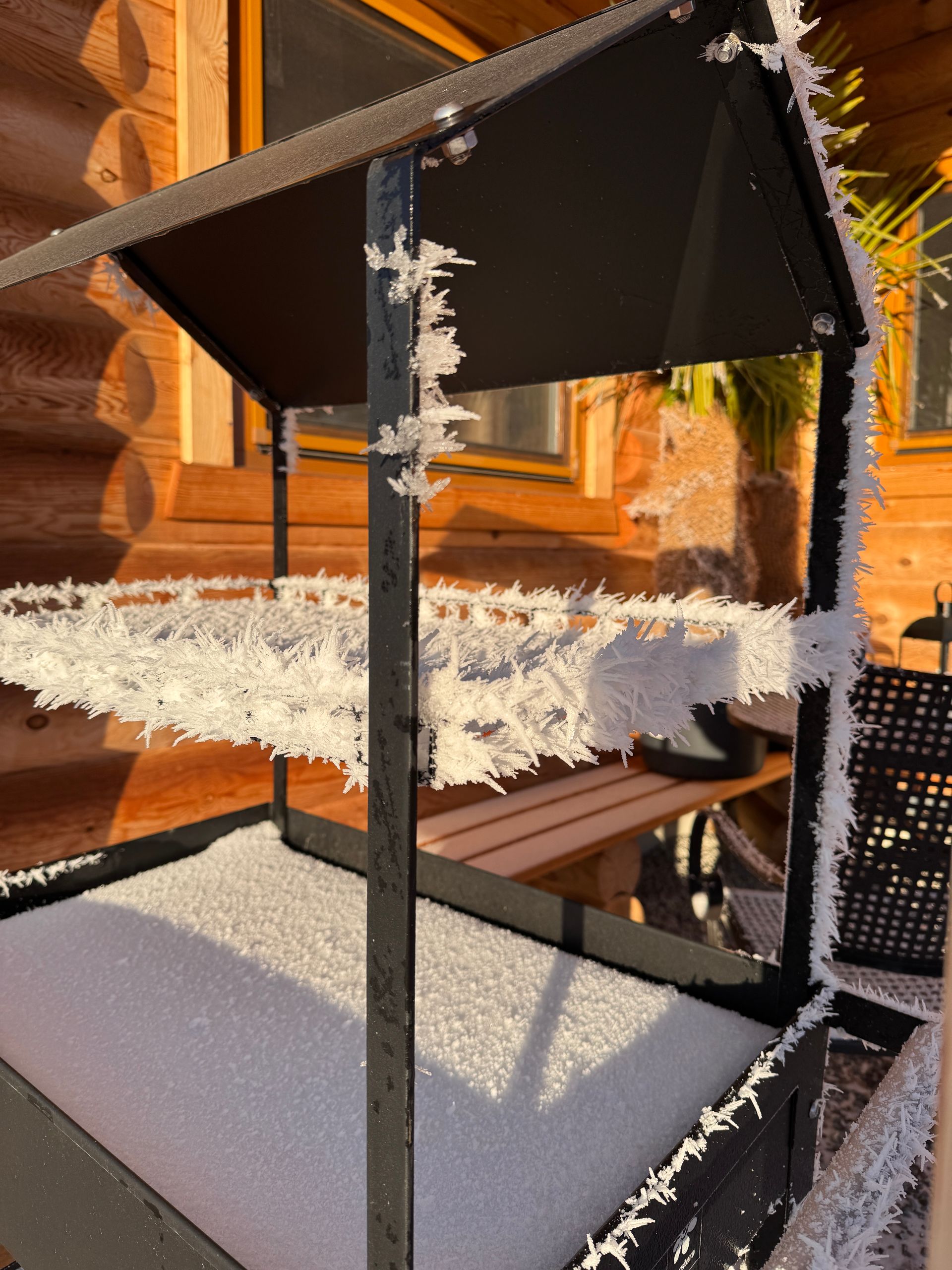 Eine schwarze, mit Schnee und Frost bedeckte Feuerstelle aus Metall steht auf einer Holzterrasse; im Hintergrund ist eine Holzhütte zu sehen.