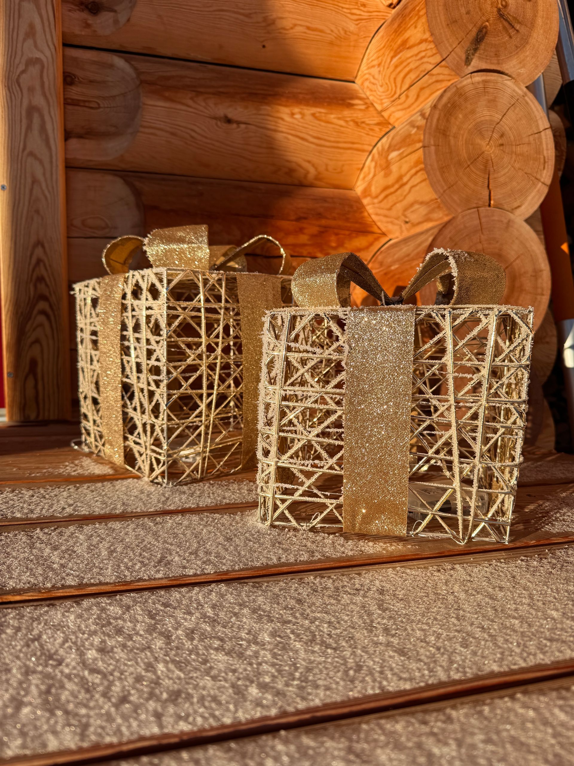 Zwei goldene, glitzernde Geschenkboxen vor einer Blockhauswand.