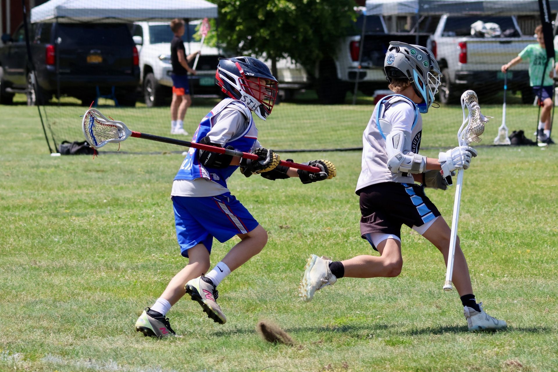 Two lacrosse players running on a grassy field, one with blue and white uniform, the other with a gray and black uniform.