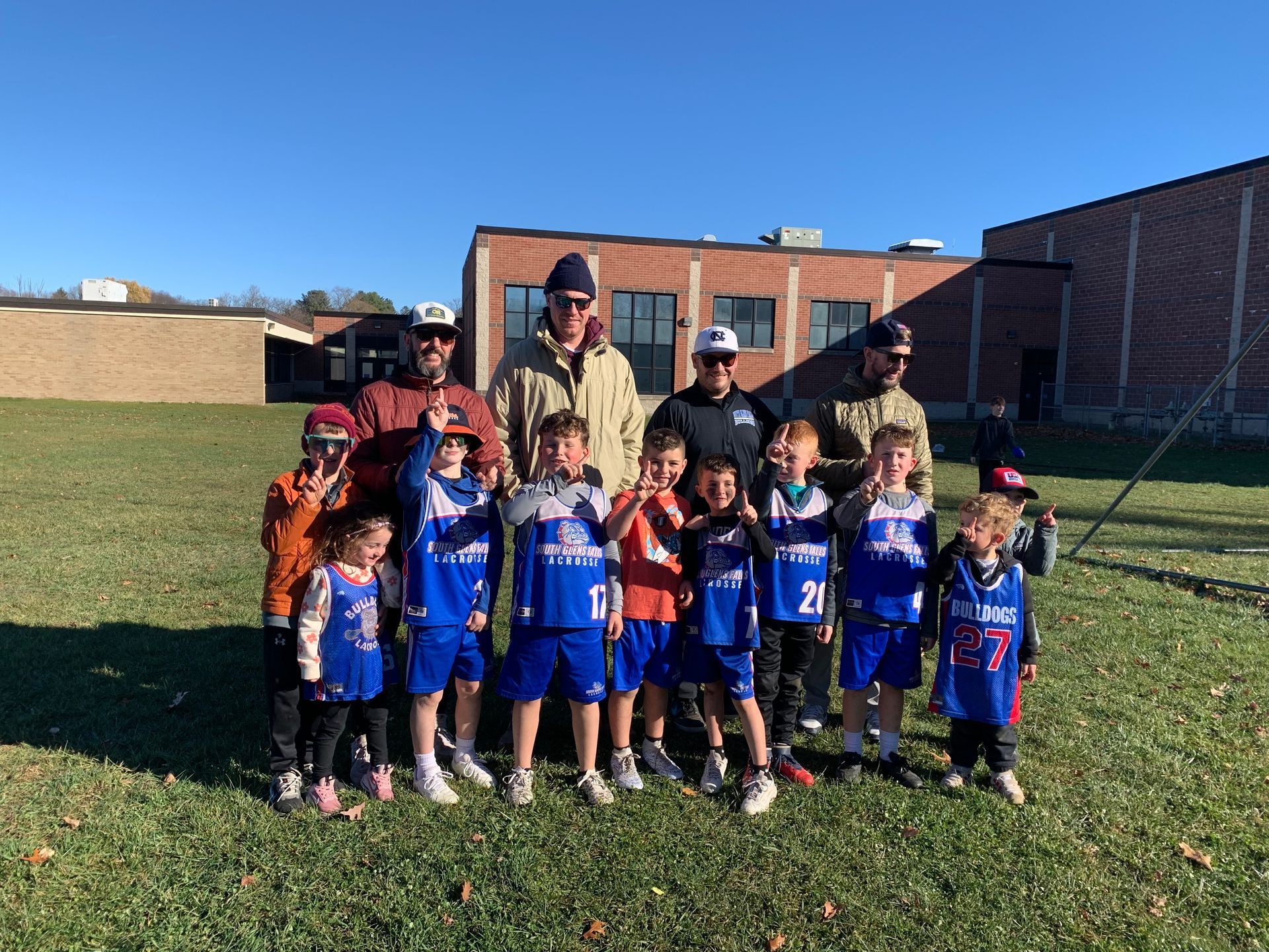 Youth basketball team in blue uniforms poses with coaches on a grassy field in front of a brick building.