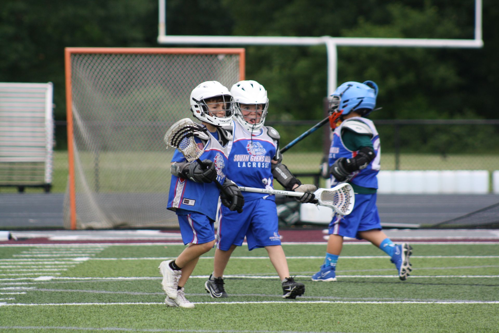 Lacrosse players in blue uniforms on a green field. Two players stand together, one with stick. A goal is in the background.