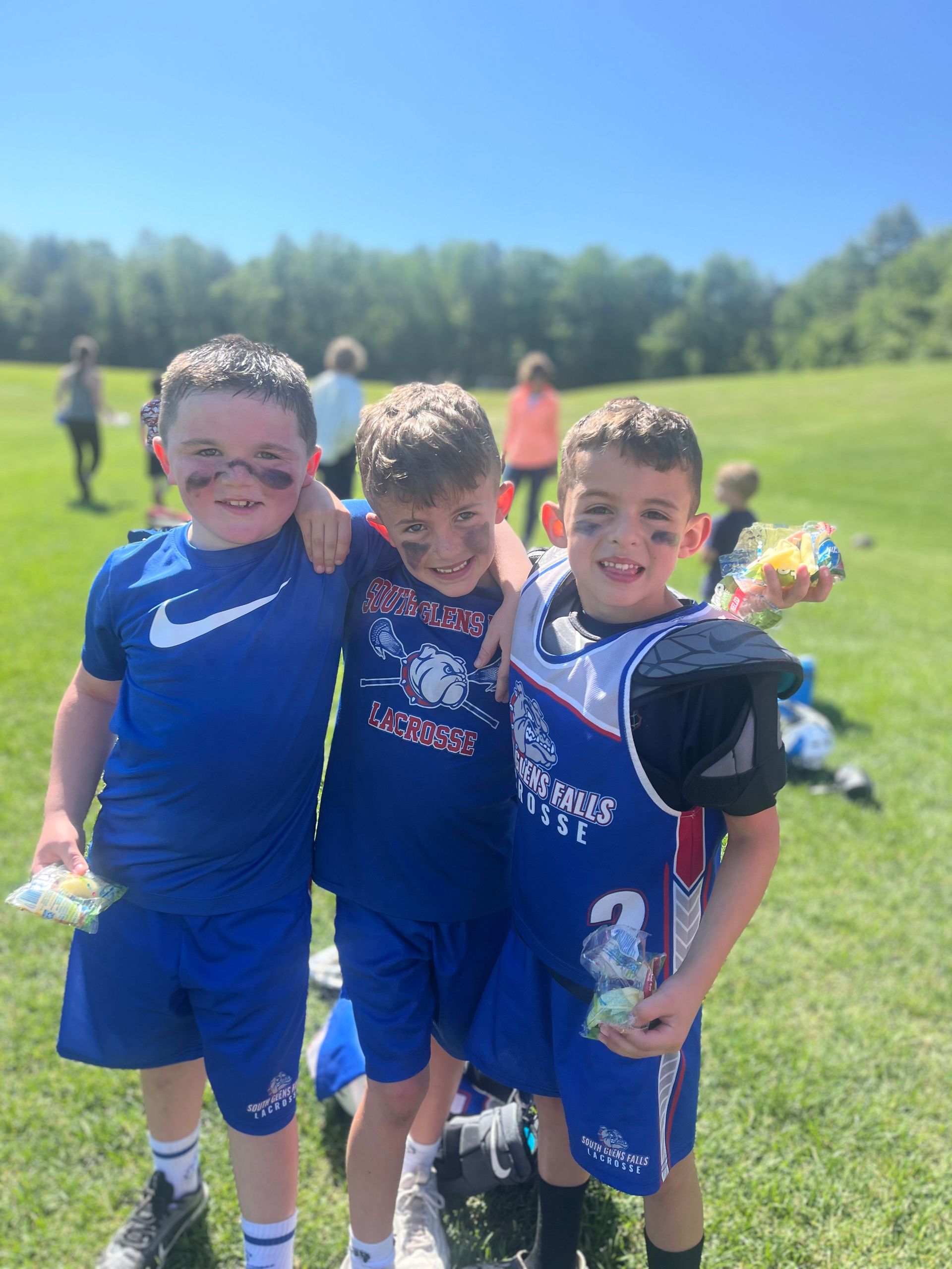 Three boys in blue sports uniforms smiling on a grassy field.