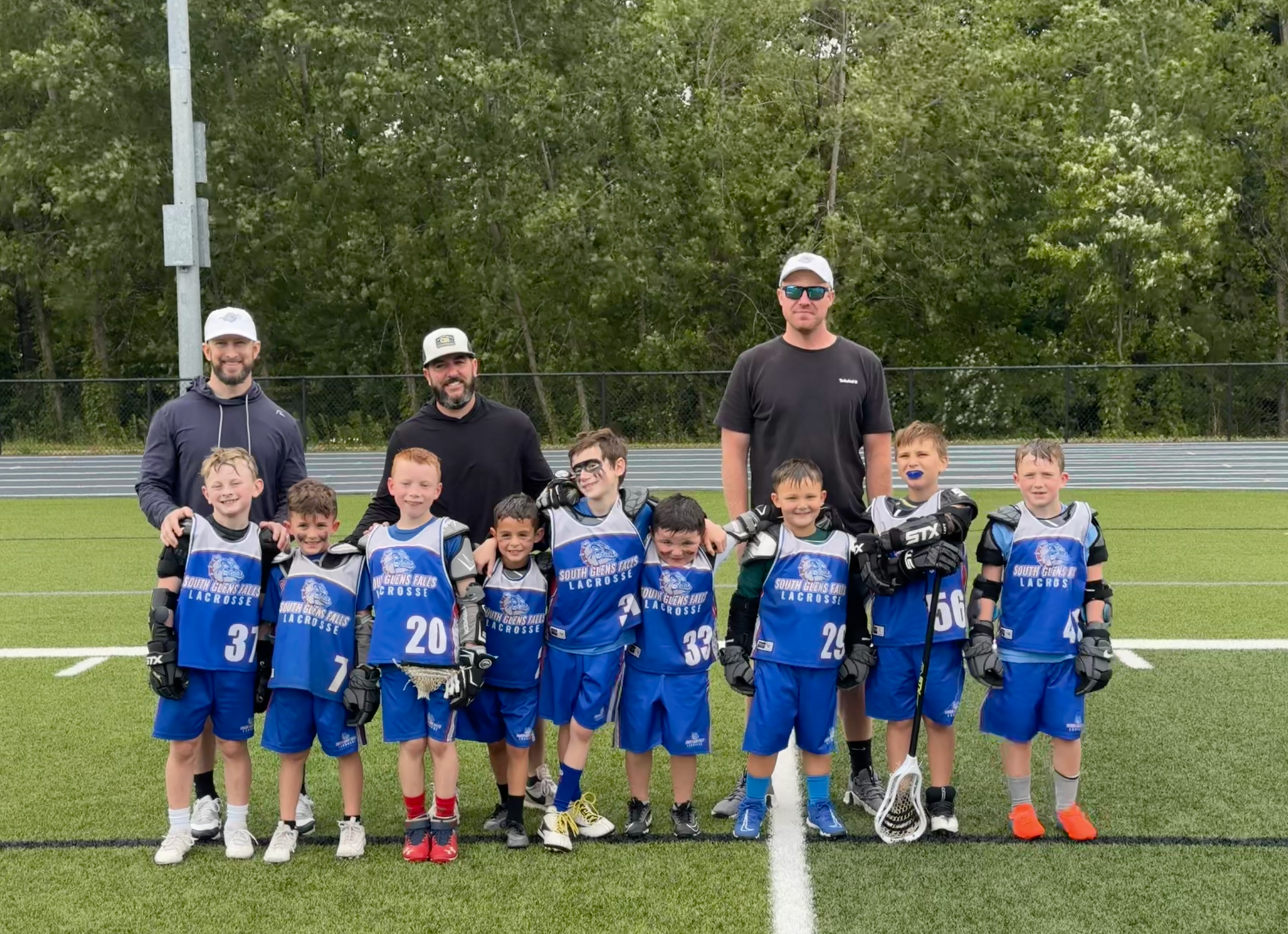 Youth lacrosse team in blue uniforms poses with coaches on a green field.