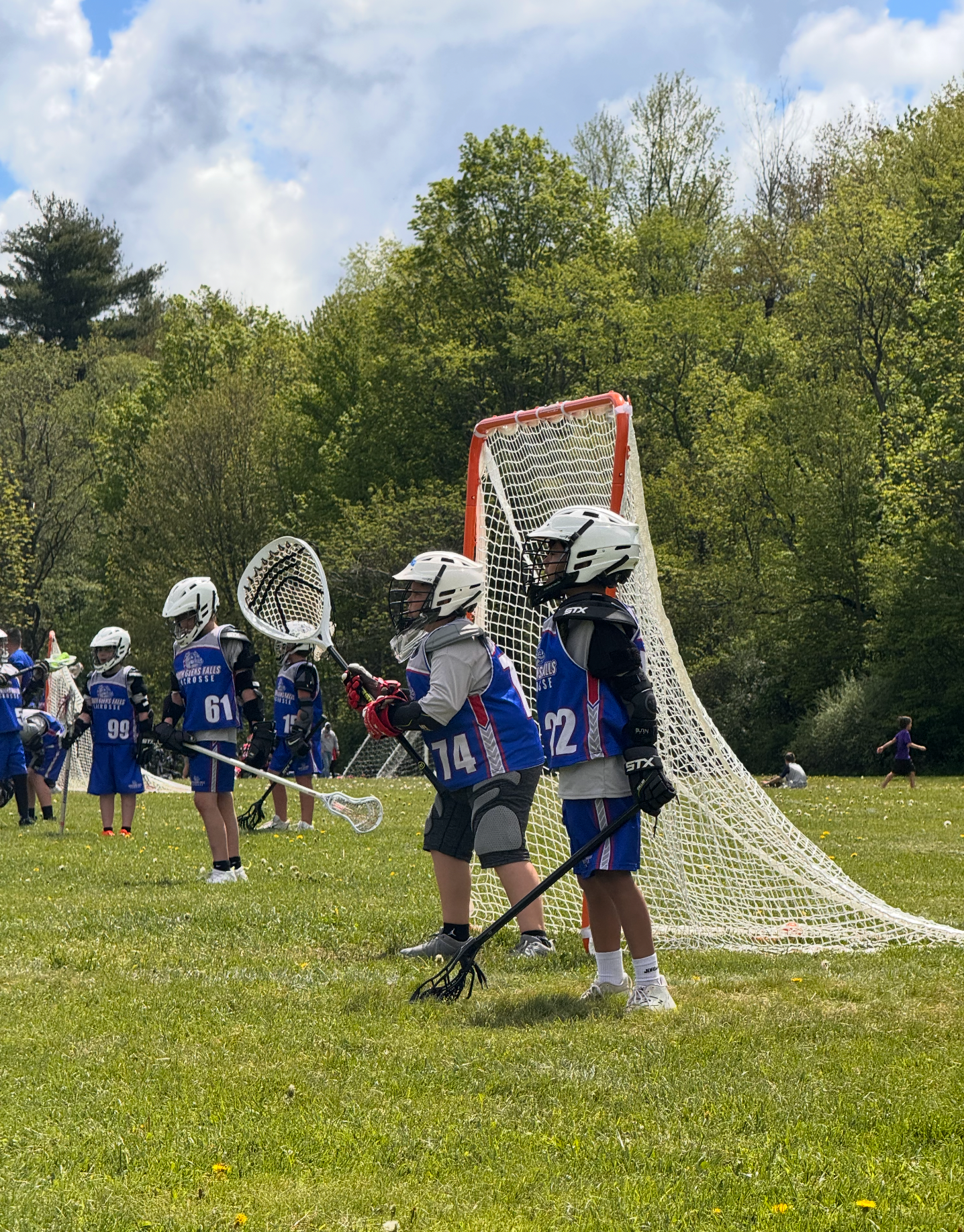 Youth lacrosse players in blue and white uniforms stand on a green field in front of a lacrosse goal.
