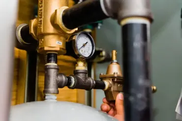 Close-up of plumbing components: brass pressure regulator, gauge, and black pipes. A hand adjusts the regulator.