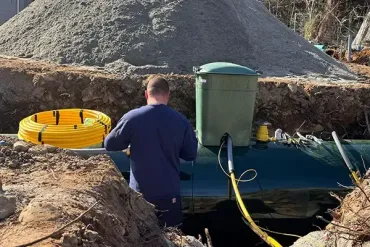 Man working on a green septic tank in a trench; yellow tubing and gravel pile nearby.