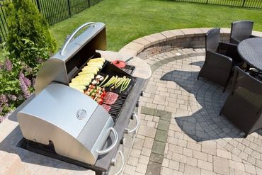 Stainless steel grill with food cooking on a stone patio, near a round table and chairs.