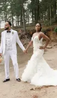 Bride and groom in white outfits holding hands outdoors in a wooded area