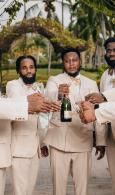 Three men in beige suits holding a bottle, standing outdoors under an archway.